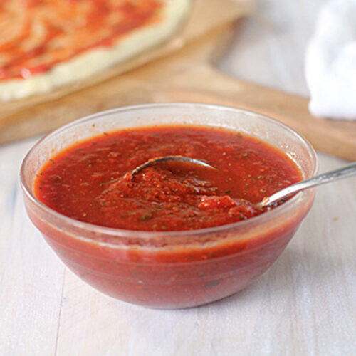 A clear glass bowl filled with red pizza sauce, with a spoon resting inside. In the background, a pizza crust being topped with sauce is visible on a wooden board.