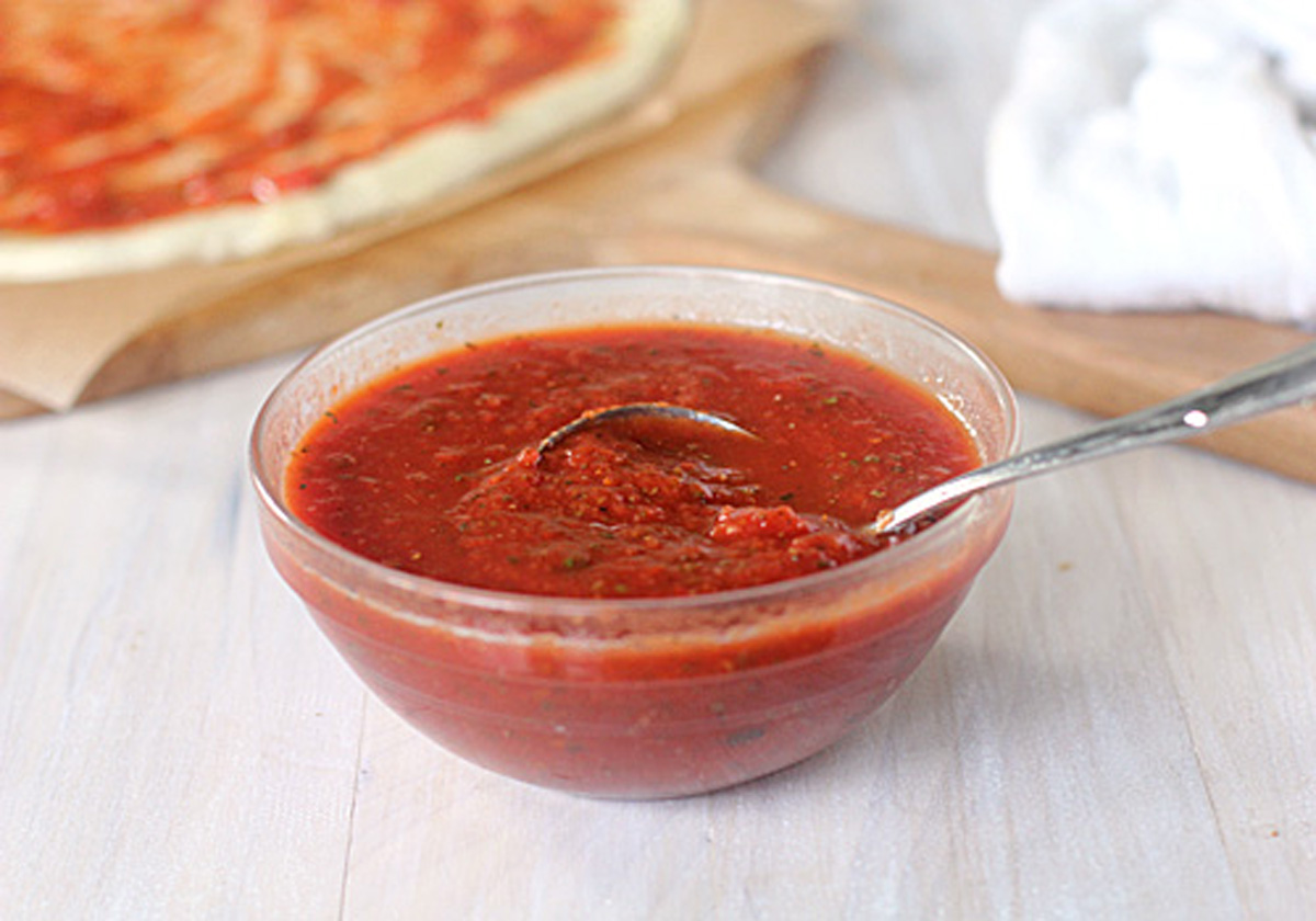 A clear glass bowl filled with red pizza sauce, with a spoon resting inside. In the background, a pizza crust being topped with sauce is visible on a wooden board.