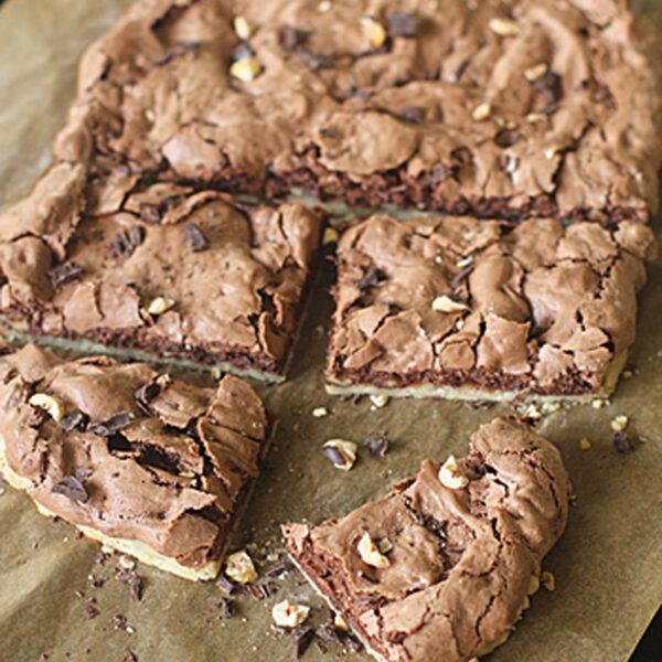 A batch of chocolate brownies with a cracked top and nut pieces is cut into squares on parchment paper, with one piece partially separated and a knife nearby.