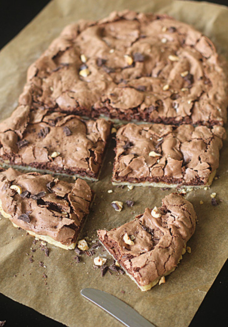A batch of chocolate brownies with a cracked top and nut pieces is cut into squares on parchment paper, with one piece partially separated and a knife nearby.