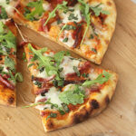 A sliced pizza on a wooden board topped with melted cheese, tomato sauce, fresh basil, and arugula leaves. One slice is slightly pulled out from the rest.