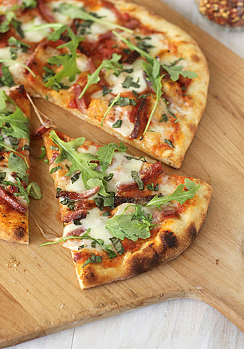A sliced pizza on a wooden board topped with melted cheese, tomato sauce, fresh basil, and arugula leaves. One slice is slightly pulled out from the rest.