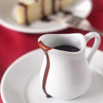 A small white pitcher of chocolate sauce with some sauce dripped down the side, placed on a white plate. In the background, a slice of cake with chocolate drizzle is on another plate, all set on a red cloth.