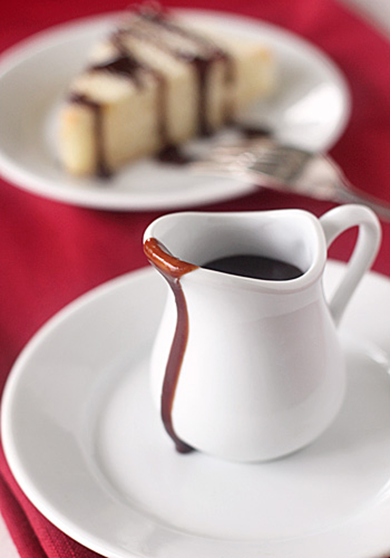 A small white pitcher of chocolate sauce with some sauce dripped down the side, placed on a white plate. In the background, a slice of cake with chocolate drizzle is on another plate, all set on a red cloth.