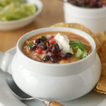 A bowl of creamy soup topped with black beans, diced tomatoes, cilantro, and a dollop of sour cream, served with tortilla chips on the side. A spoon rests on the plate, with lime wedges and salsa in the background.