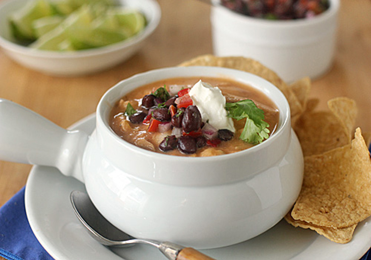 A bowl of creamy soup topped with black beans, diced tomatoes, cilantro, and a dollop of sour cream, served with tortilla chips on the side. A spoon rests on the plate, with lime wedges and salsa in the background.