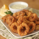 A plate of crispy fried calamari rings served with lemon wedges, parsley, and a small bowl of creamy dipping sauce on a light-colored napkin.