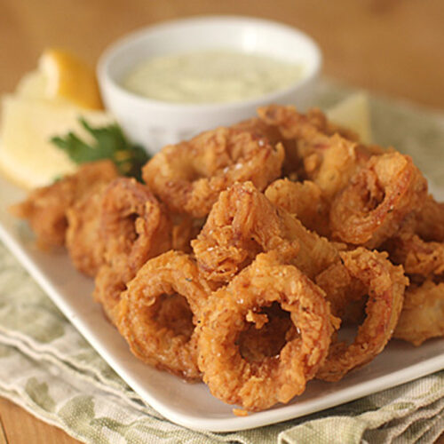 A plate of crispy fried calamari rings served with lemon wedges, parsley, and a small bowl of creamy dipping sauce on a light-colored napkin.