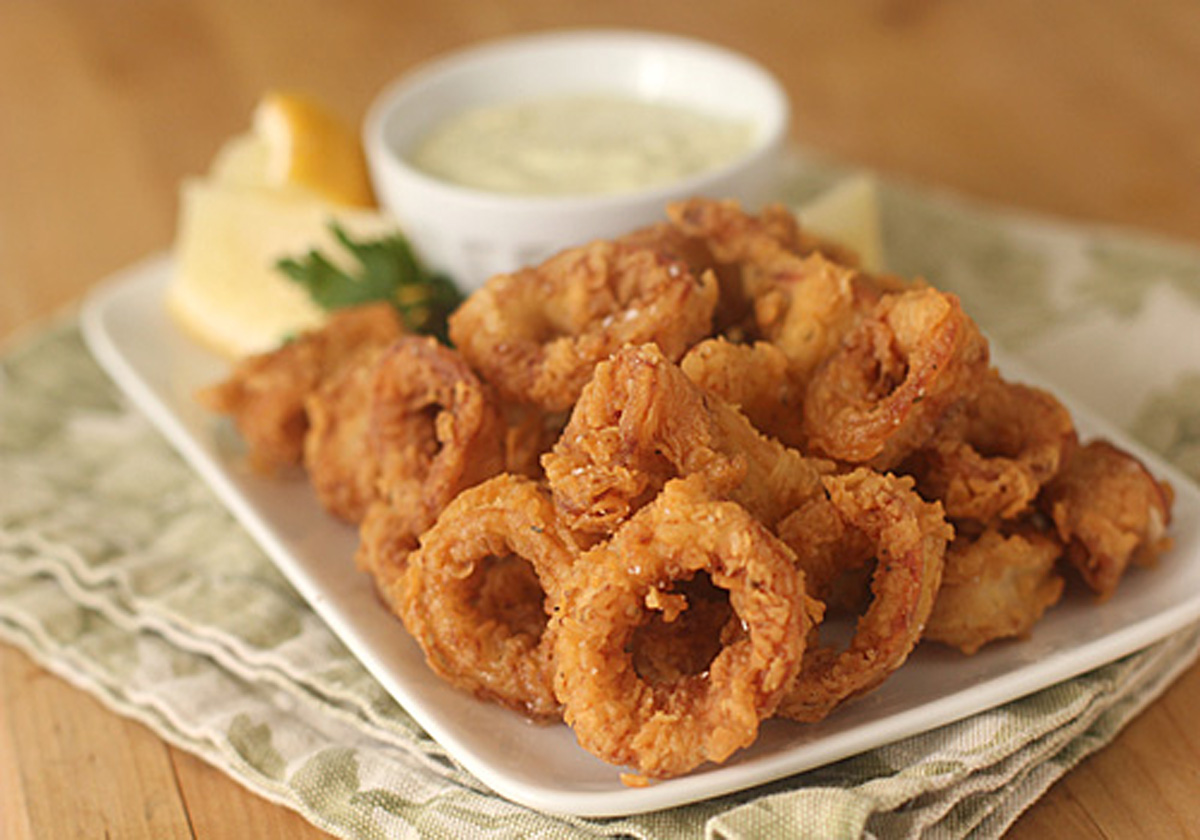 A plate of crispy fried calamari rings served with lemon wedges, parsley, and a small bowl of creamy dipping sauce on a light-colored napkin.