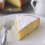 A slice of yellow cake dusted with powdered sugar sits on a white plate with a fork. A small pitcher and the rest of the cake on a platter are in the background.