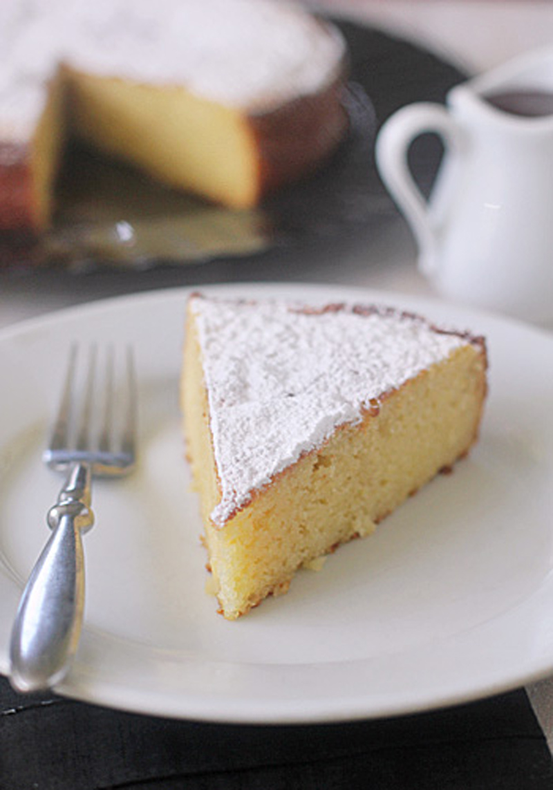 A slice of yellow cake dusted with powdered sugar sits on a white plate with a fork. A small pitcher and the rest of the cake on a platter are in the background.