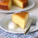 A slice of yellow cake on a white plate with a dollop of cream, next to a fork, with the rest of the cake in the background. The plate sits on a folded blue napkin.