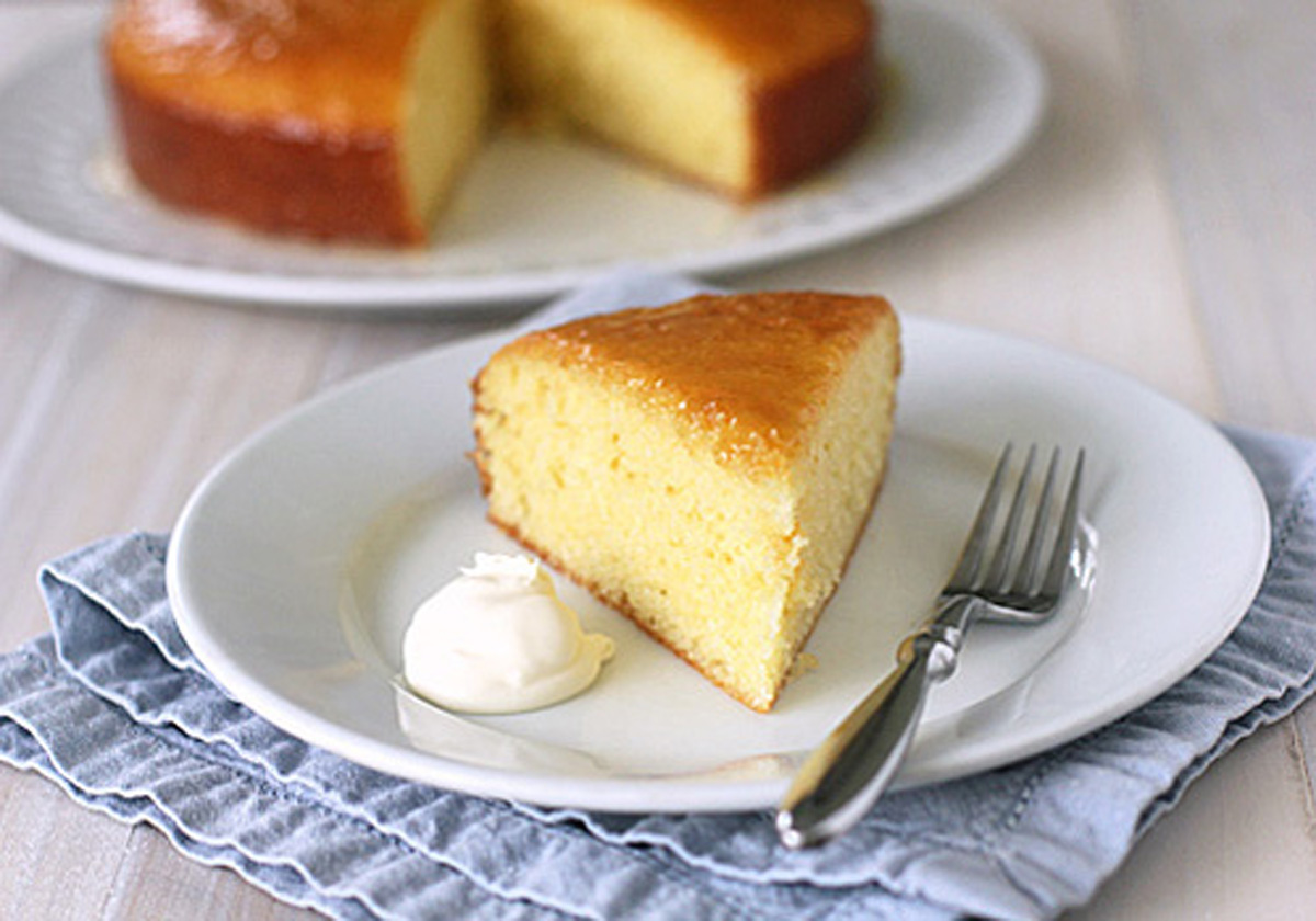 A slice of yellow cake on a white plate with a dollop of cream, next to a fork, with the rest of the cake in the background. The plate sits on a folded blue napkin.