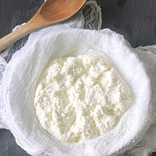 A bowl lined with cheesecloth holds fresh, crumbly homemade ricotta cheese, with a wooden spoon resting nearby on a dark surface.