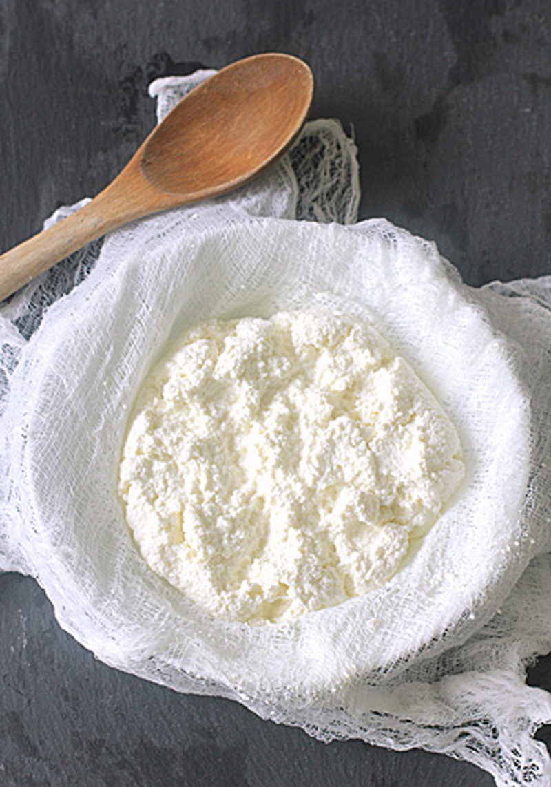 A bowl lined with cheesecloth holds fresh, crumbly homemade ricotta cheese, with a wooden spoon resting nearby on a dark surface.