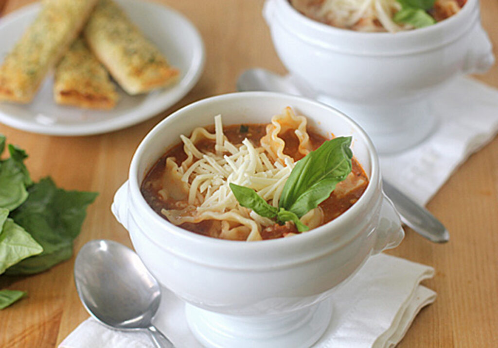 A white bowl filled with lasagna soup, topped with shredded cheese and fresh basil, sits on a napkin with a spoon beside it. Breadsticks and basil leaves are visible in the background.