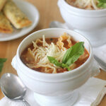 A white bowl filled with lasagna soup, topped with shredded cheese and fresh basil, sits on a napkin with a spoon beside it. Breadsticks and basil leaves are visible in the background.