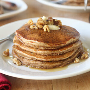 A stack of pancakes topped with butter, chopped walnuts, and syrup sits on a white plate with a fork, next to a red napkin on a wooden table.