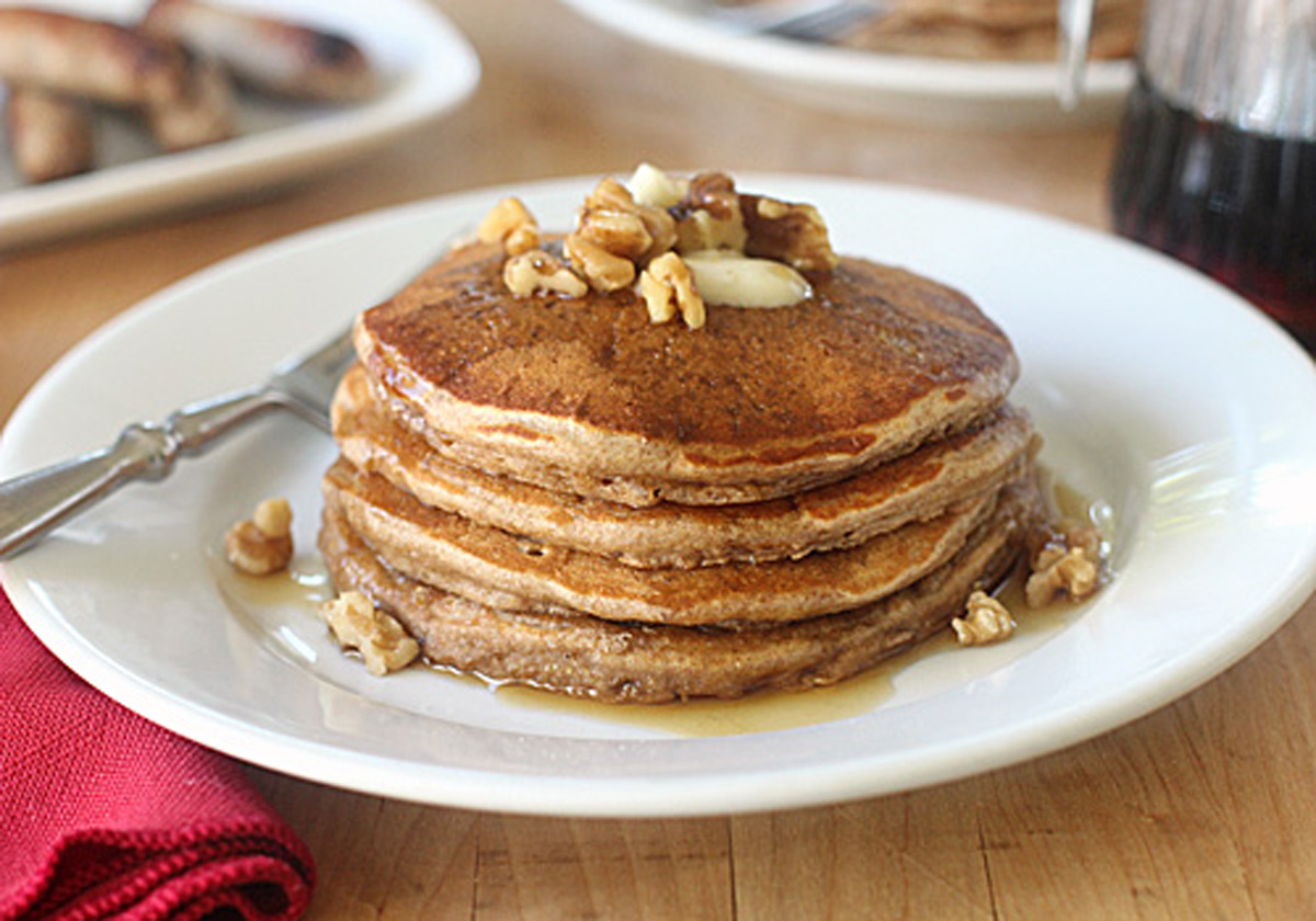 A stack of pancakes topped with butter, chopped walnuts, and syrup sits on a white plate with a fork, next to a red napkin on a wooden table.