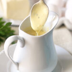 A spoon drizzles creamy yellow sauce, possibly hollandaise, into a white ceramic pitcher on a matching saucer. Butter, eggs, and parsley are blurred in the background.