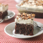 A slice of chocolate cake with whipped cream topping and chopped nuts sits on a white plate with a fork; the rest of the cake is in a glass dish in the background on a red checkered cloth.