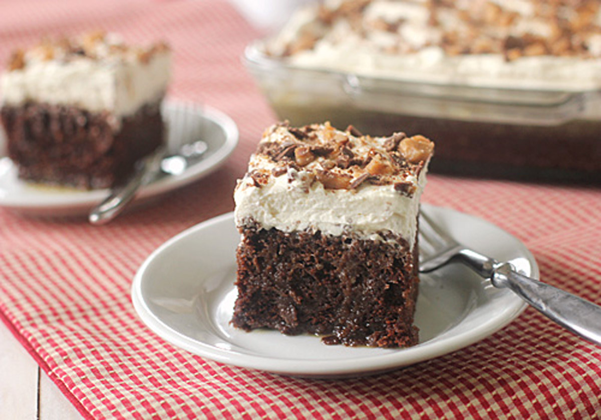 A slice of chocolate cake with whipped cream topping and chopped nuts sits on a white plate with a fork; the rest of the cake is in a glass dish in the background on a red checkered cloth.