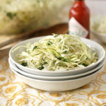 A bowl of shredded cabbage slaw sits on stacked white plates with a bottle of hot sauce and a fork in the background, atop a yellow-patterned tablecloth.