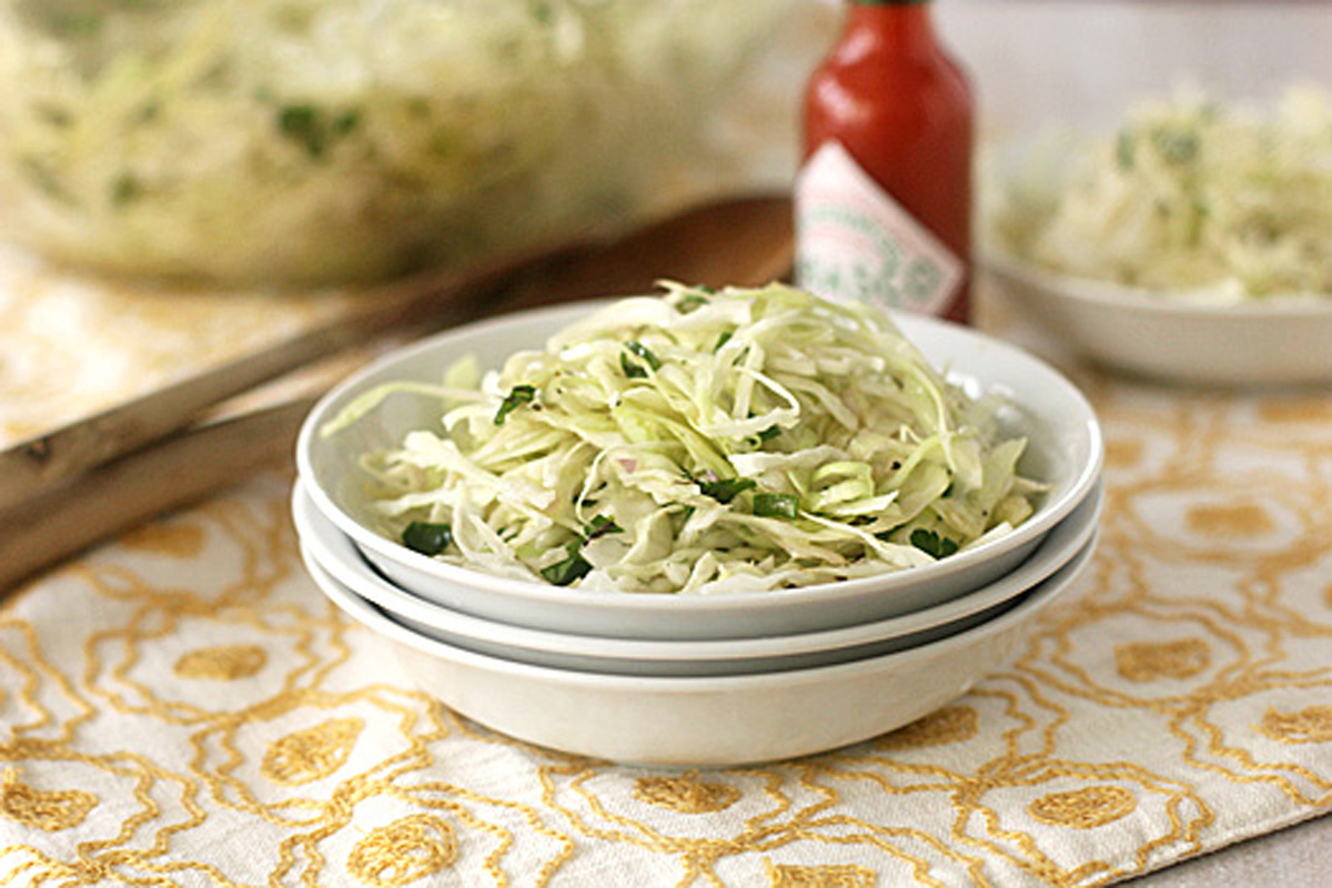 A bowl of shredded cabbage slaw sits on stacked white plates with a bottle of hot sauce and a fork in the background, atop a yellow-patterned tablecloth.