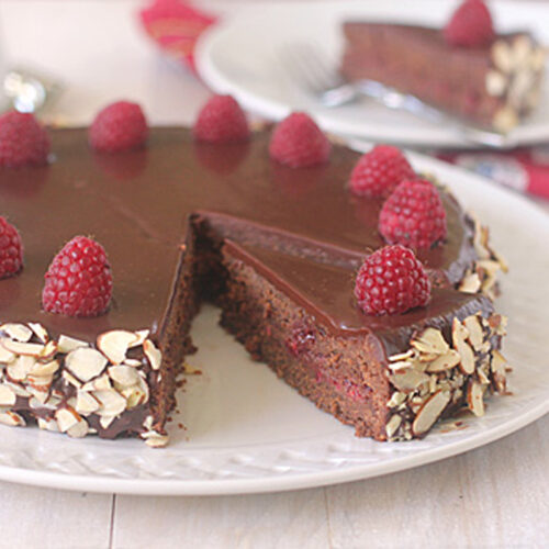 A chocolate cake topped with raspberries and coated in a glossy chocolate glaze sits on a white plate. Sliced almonds decorate the sides. A slice is missing, and it rests on a plate in the background.