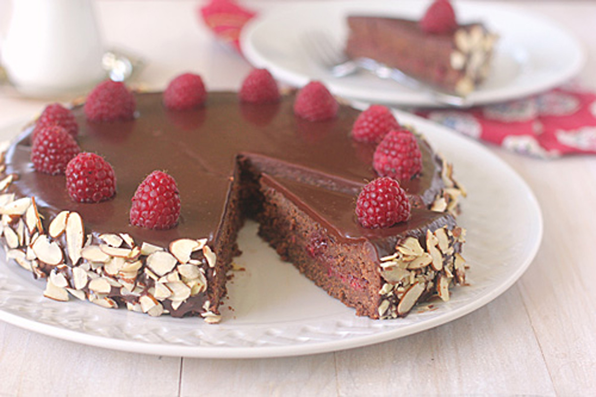 A chocolate cake topped with raspberries and coated in a glossy chocolate glaze sits on a white plate. Sliced almonds decorate the sides. A slice is missing, and it rests on a plate in the background.
