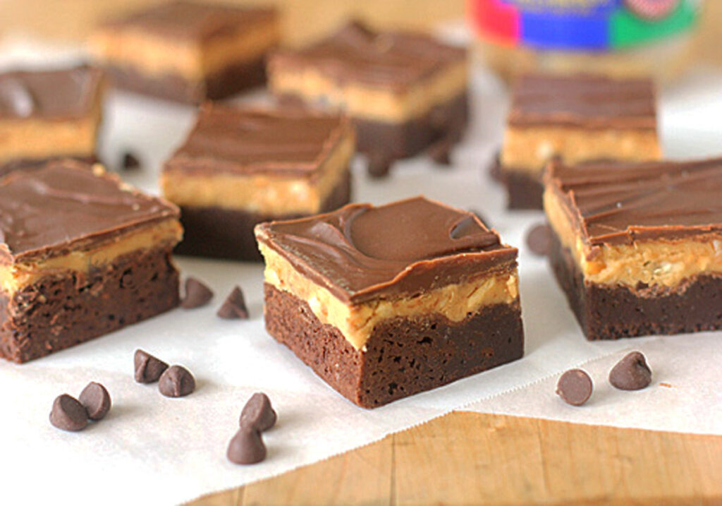 Chocolate and peanut butter layered dessert bars are arranged on parchment paper with a few chocolate chips scattered around. Each bar has a brownie base, a peanut butter middle layer, and a chocolate top layer.