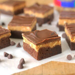 Chocolate and peanut butter layered dessert bars are arranged on parchment paper with a few chocolate chips scattered around. Each bar has a brownie base, a peanut butter middle layer, and a chocolate top layer.