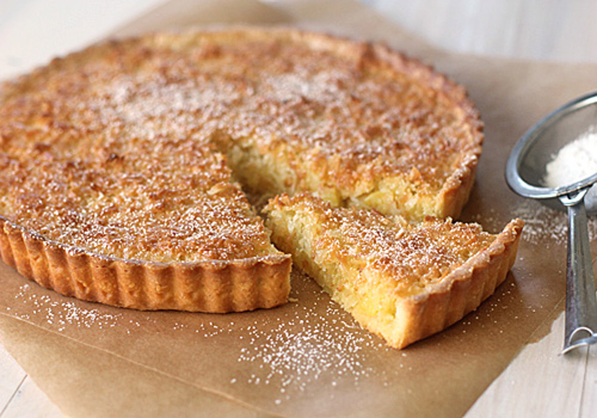 A golden-brown tart with a slice cut out sits on brown parchment paper. The tart is dusted with powdered sugar, and a metal sieve with powdered sugar is nearby.