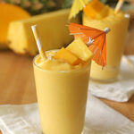 Two glasses of creamy yellow smoothie garnished with pineapple and mango chunks, striped straws, and colorful paper umbrellas, set on white napkins with fresh pineapple and mango in the background.