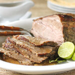 A white plate with sliced roasted beef, lime wedges, and a serving fork, with a larger piece of roast beef and a gravy boat in the background.