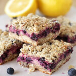 Three blueberry crumb bars are stacked on parchment paper, with a few loose blueberries nearby. In the background, there are two yellow lemons, one of which is cut in half. The bars have a crumbly oat topping.