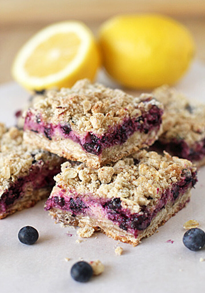 Three blueberry crumb bars are stacked on parchment paper, with a few loose blueberries nearby. In the background, there are two yellow lemons, one of which is cut in half. The bars have a crumbly oat topping.