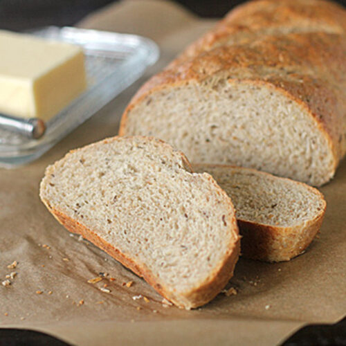 A loaf of sliced bread on brown parchment paper, with two slices in the foreground and a glass butter dish with a stick of butter and a butter knife in the background.