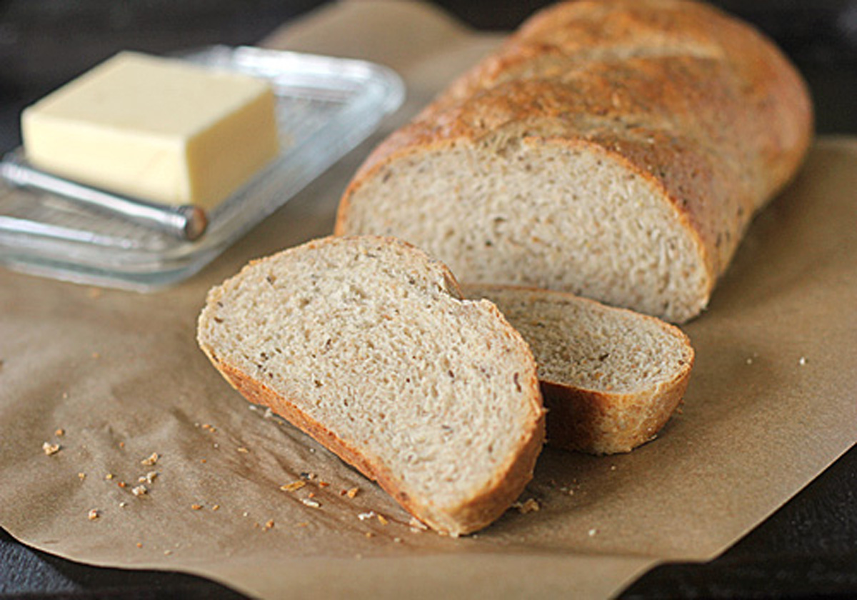 A loaf of sliced bread on brown parchment paper, with two slices in the foreground and a glass butter dish with a stick of butter and a butter knife in the background.