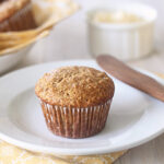 A bran muffin sits on a white plate with a wooden knife, next to a yellow napkin. In the background, there is a bowl of more muffins, a ramekin with spread, and a glass of orange juice.