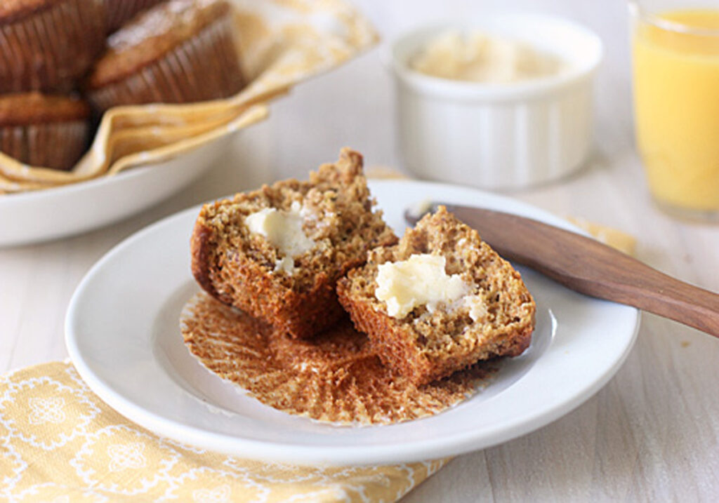 A bran muffin split in half with butter melting on top, served on a white plate with a wooden knife. In the background, there’s a bowl of more muffins, a ramekin of butter, and a glass of orange juice.