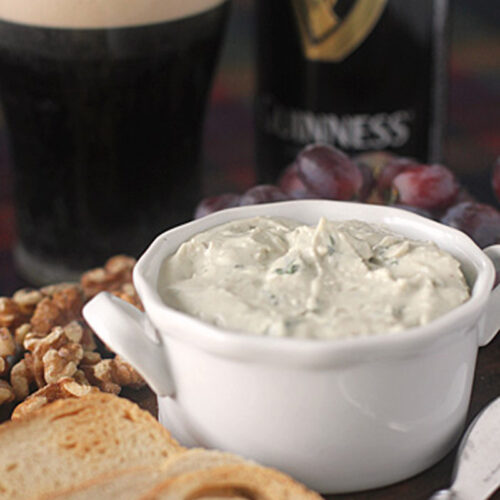 A white bowl of creamy cheese dip sits on a wooden board with slices of bread, walnuts, and grapes. In the background, there is a glass and a bottle of Guinness beer.