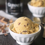 A scoop of light brown ice cream with chocolate chunks in a white bowl, with another bowl and a blurred can in the background. Some chocolate pieces and two spoons are nearby on a dark surface.