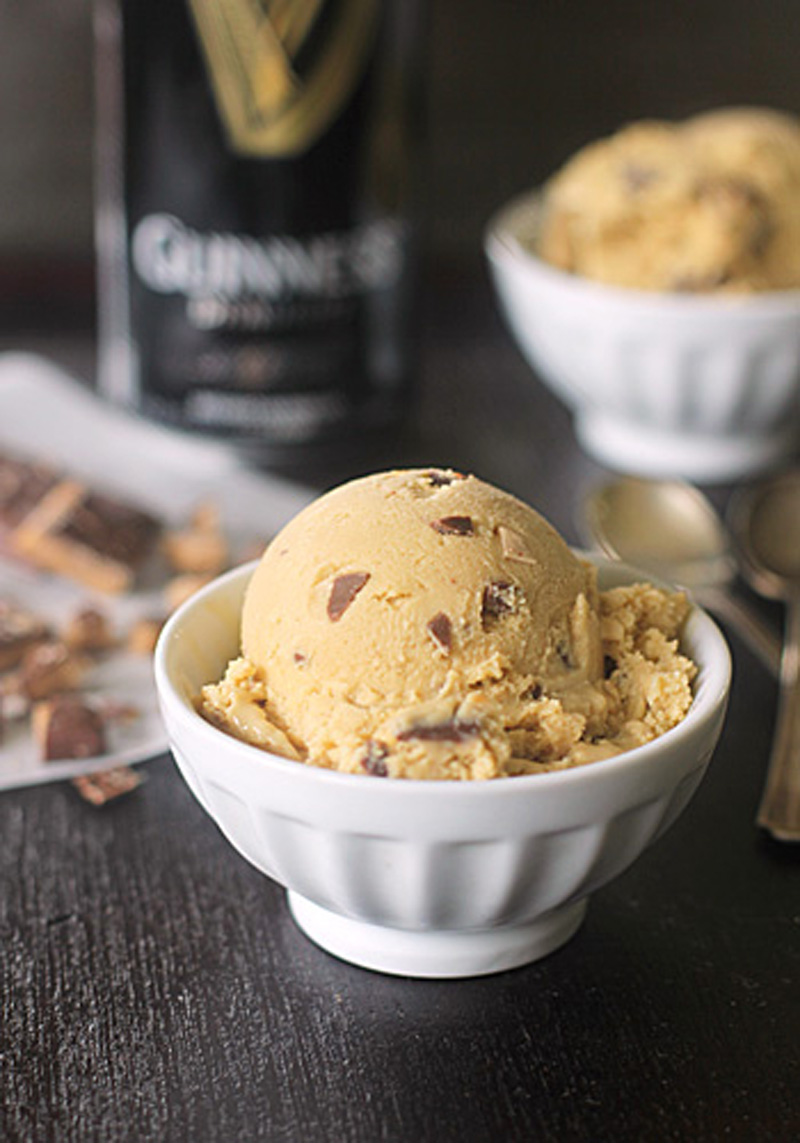 A scoop of light brown ice cream with chocolate chunks in a white bowl, with another bowl and a blurred can in the background. Some chocolate pieces and two spoons are nearby on a dark surface.