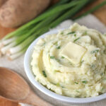 A bowl of mashed potatoes with chopped green onions and a pat of butter on top, placed on a cloth next to a wooden spoon, whole potatoes, and fresh green onions.