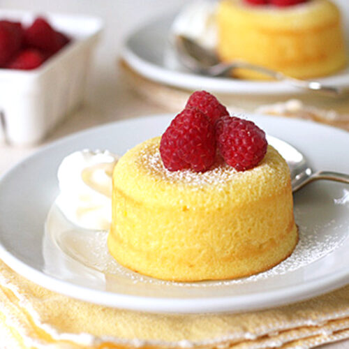 A small yellow sponge cake topped with three raspberries and dusted with powdered sugar sits on a white plate with a swirl of whipped cream, next to a spoon. A basket of raspberries is in the background.
