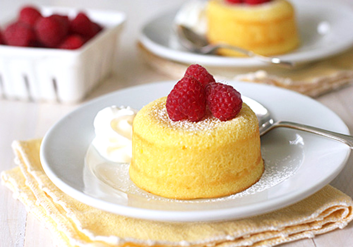 A small yellow sponge cake topped with three raspberries and dusted with powdered sugar sits on a white plate with a swirl of whipped cream, next to a spoon. A basket of raspberries is in the background.