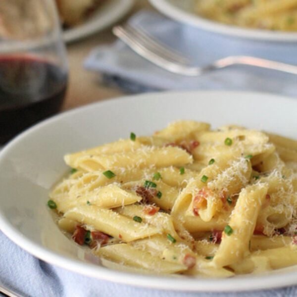 A white bowl of penne pasta with grated cheese and herbs sits on a blue napkin, with a fork nearby and a glass of red wine and bread in the background.