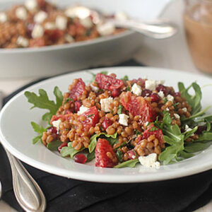 A white plate of salad with arugula, wheat berries, orange slices, pomegranate seeds, and feta cheese sits on a black napkin beside silver cutlery; a serving dish and dressing are in the background.