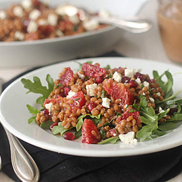 A white plate of salad with arugula, wheat berries, orange slices, pomegranate seeds, and feta cheese sits on a black napkin beside silver cutlery; a serving dish and dressing are in the background.